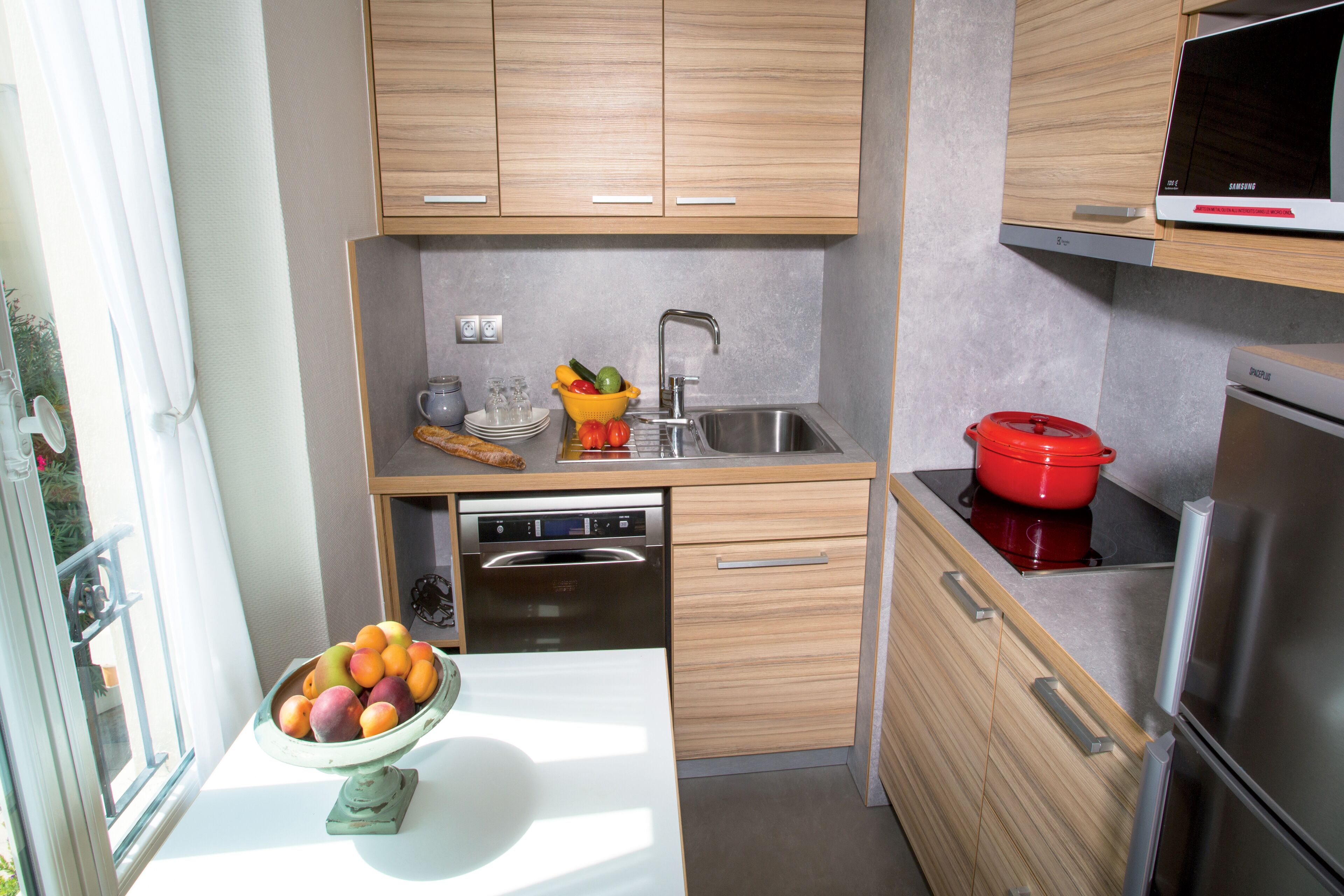 Una cocina compacta con armarios de madera, un lavamanos de acero inoxidable, una olla roja y un cuenco de fruta en la encimera.