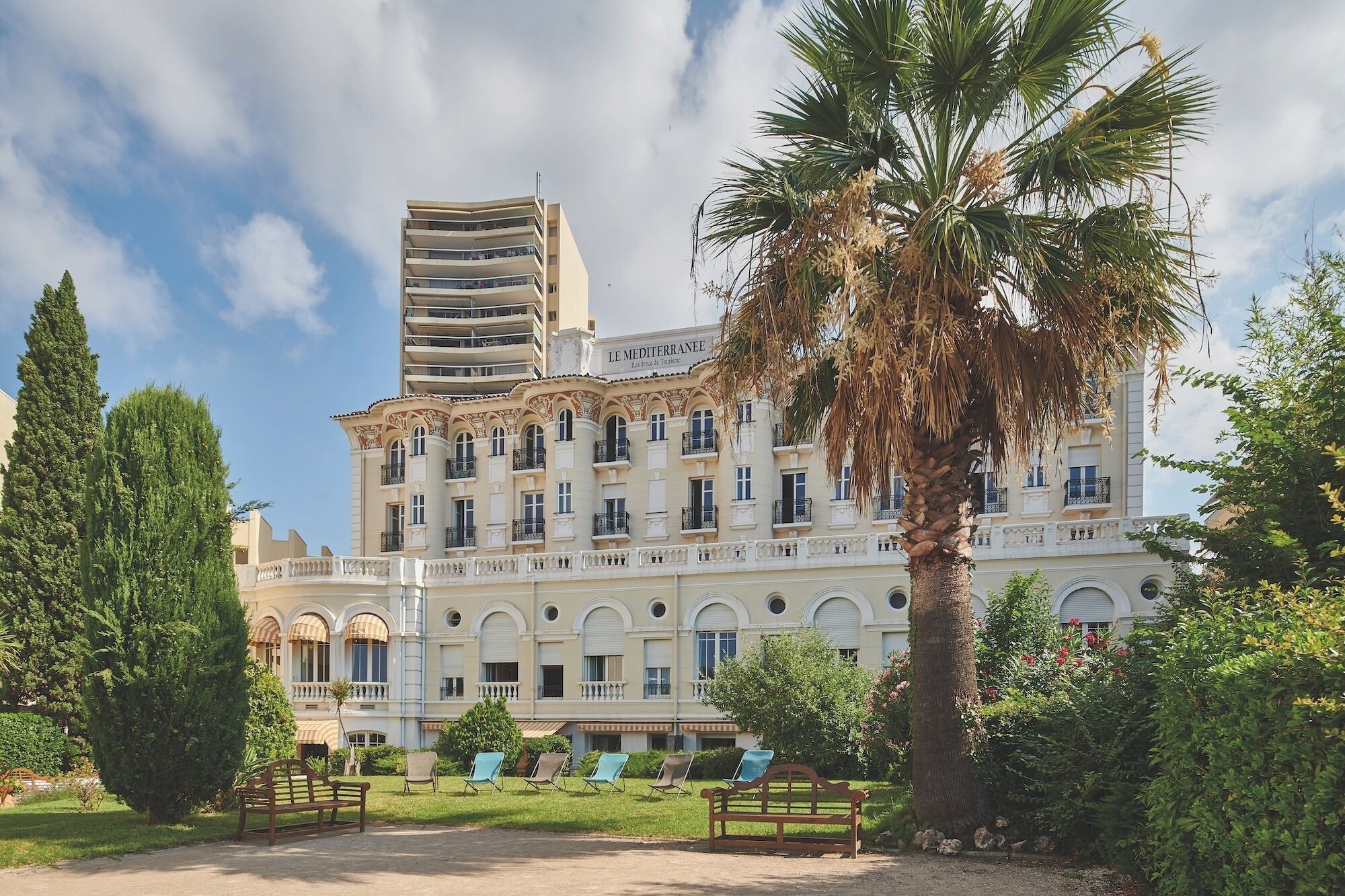 A large hotel with multiple balconies, a garden with benches, and palm trees.