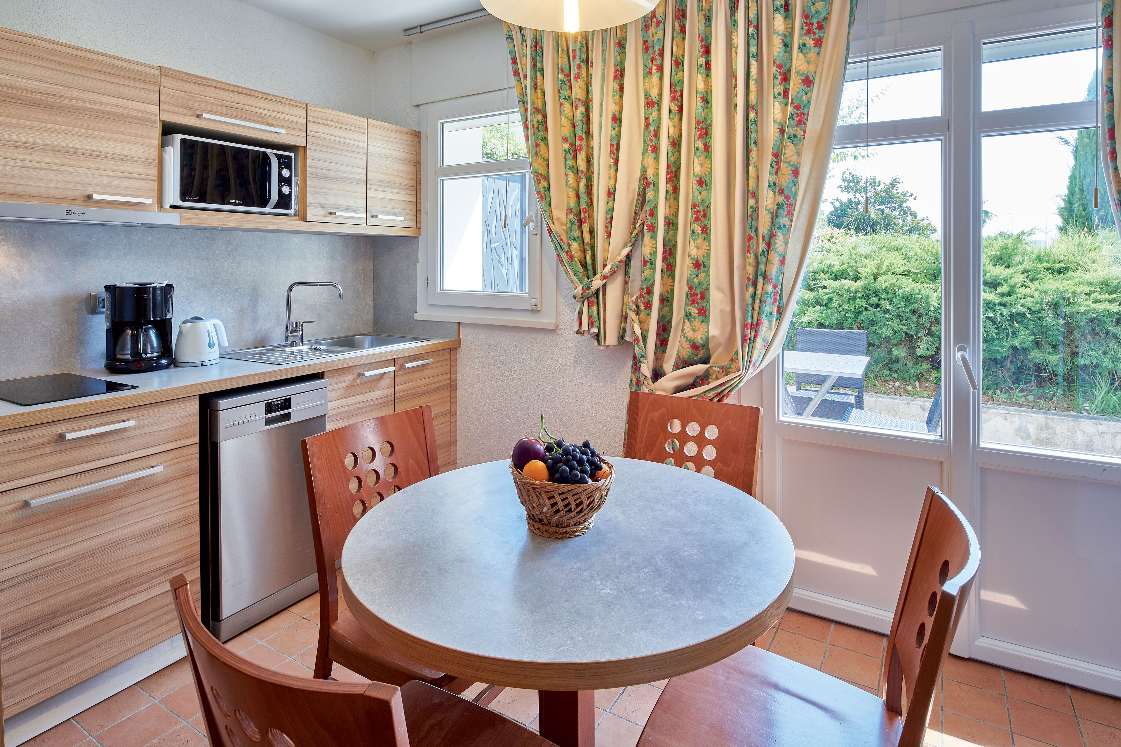 A kitchen with wooden cabinets, a microwave, a coffee maker, and a dishwasher. There is a round table with chairs and a fruit basket on it.