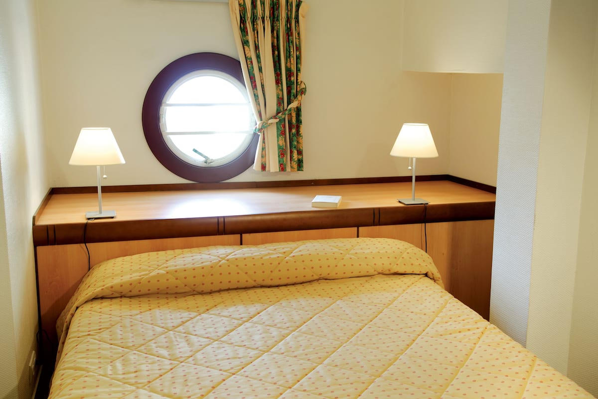 A bed with a quilted yellow bedspread, two bedside lamps, a round window with curtains, and a wooden headboard.