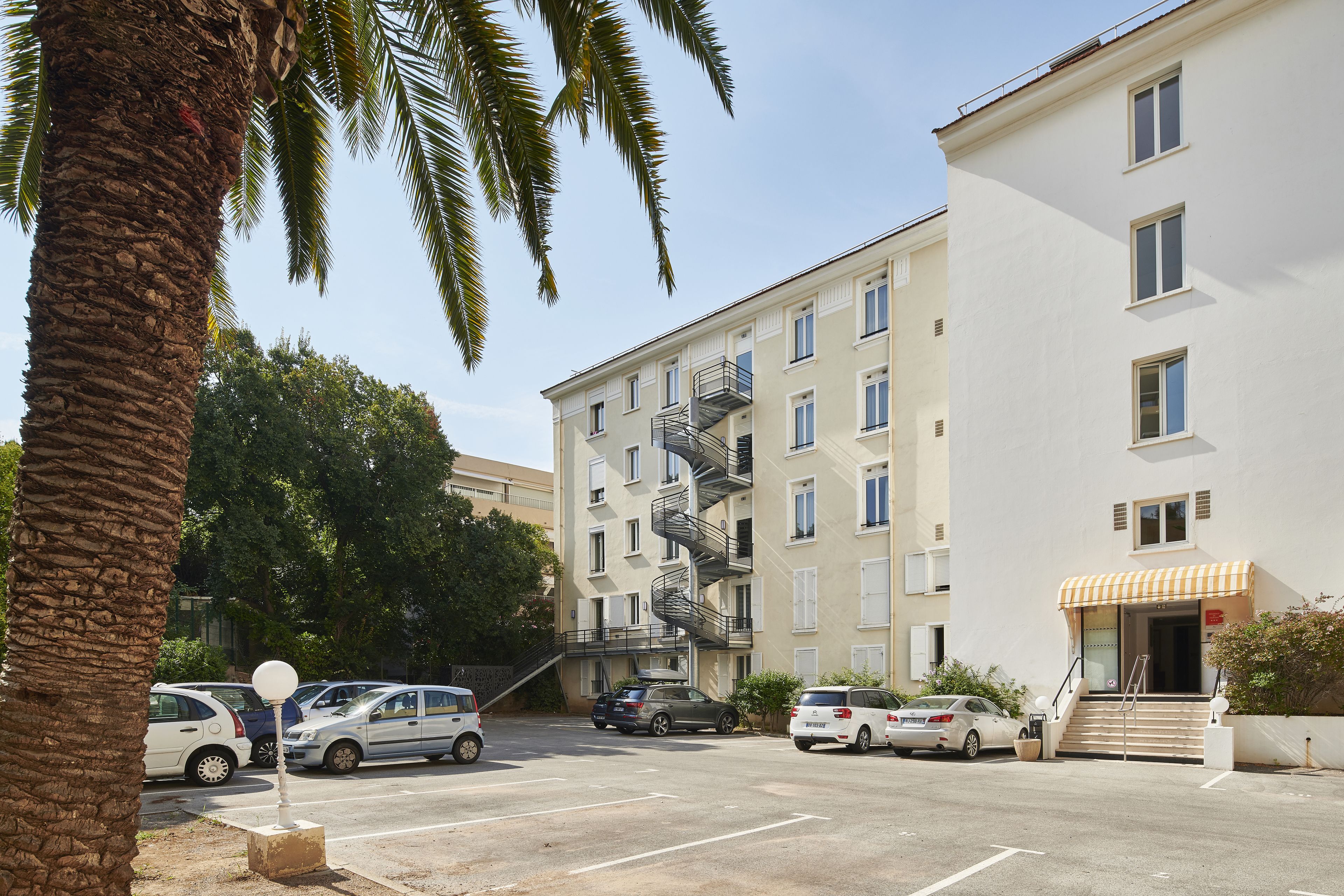 A multi-story residential building with a parking lot, a staircase leading to the entrance, and a small awning over the door.