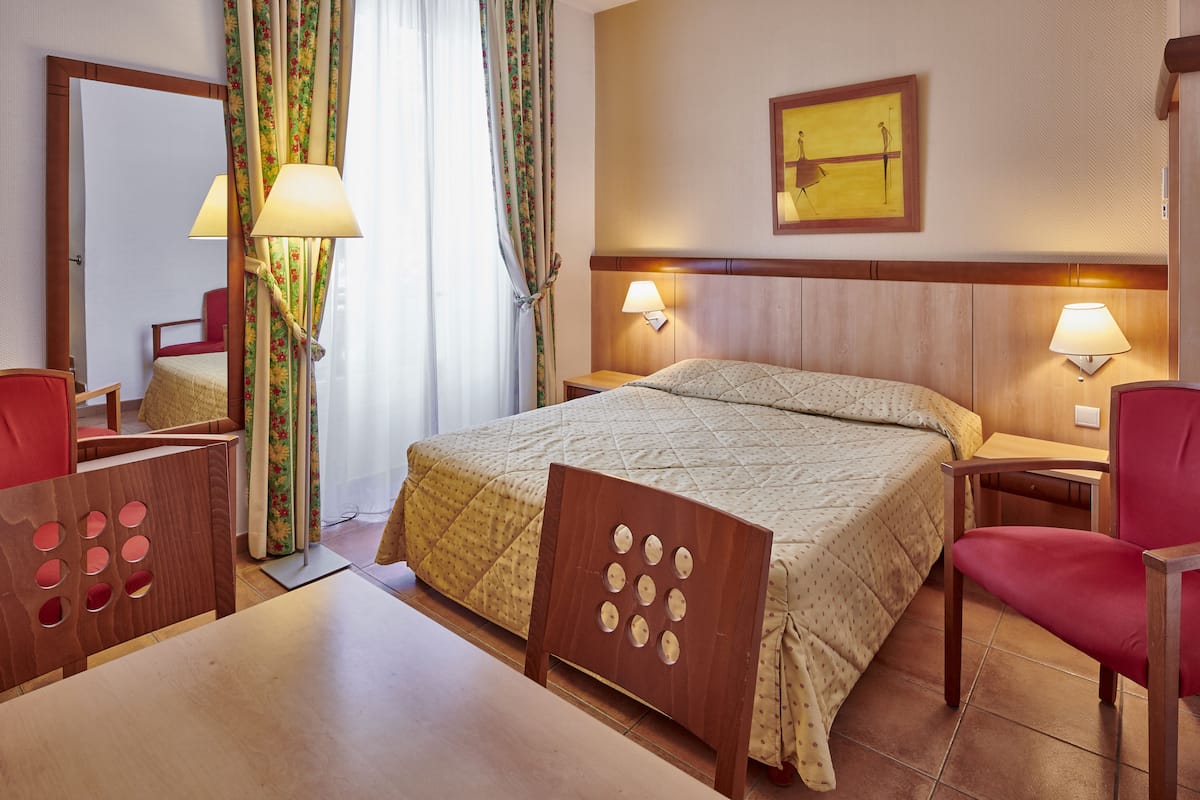 A hotel room with a bed, two bedside tables with lamps, a chair, a desk, a mirror, and a painting on the wall.