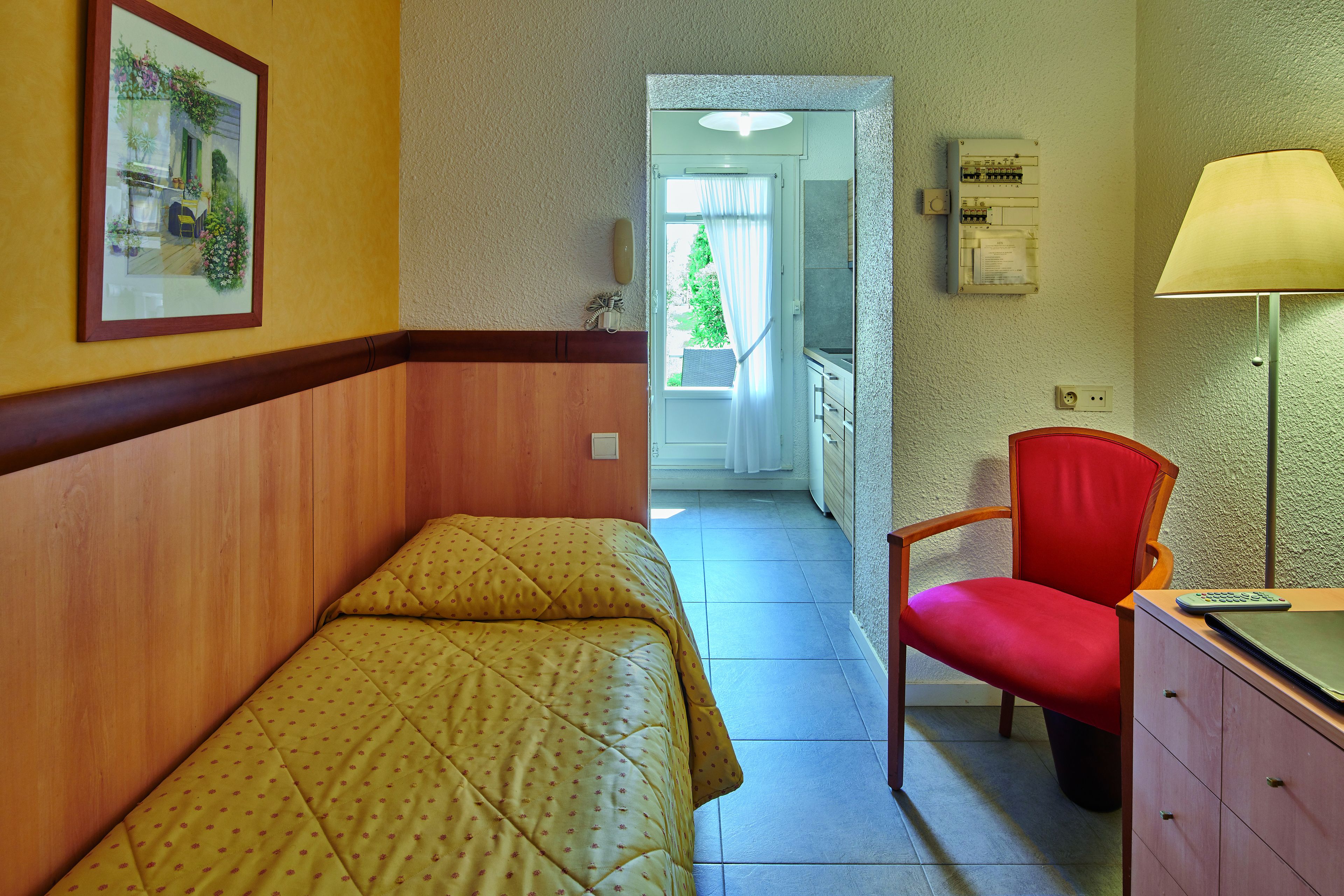 A hotel room with two single beds, a red chair, a desk with a lamp, and a framed picture on the wall.