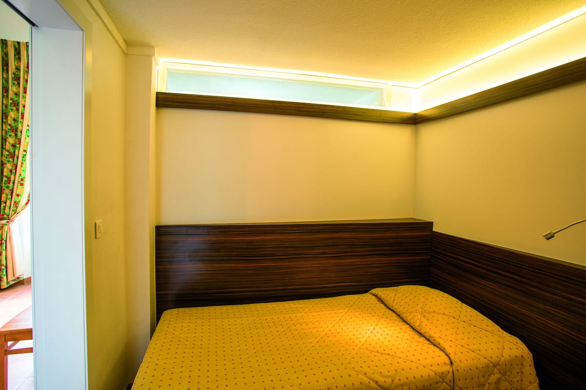 A small, neatly arranged bedroom with a single bed, a window, and a wall-mounted light fixture.