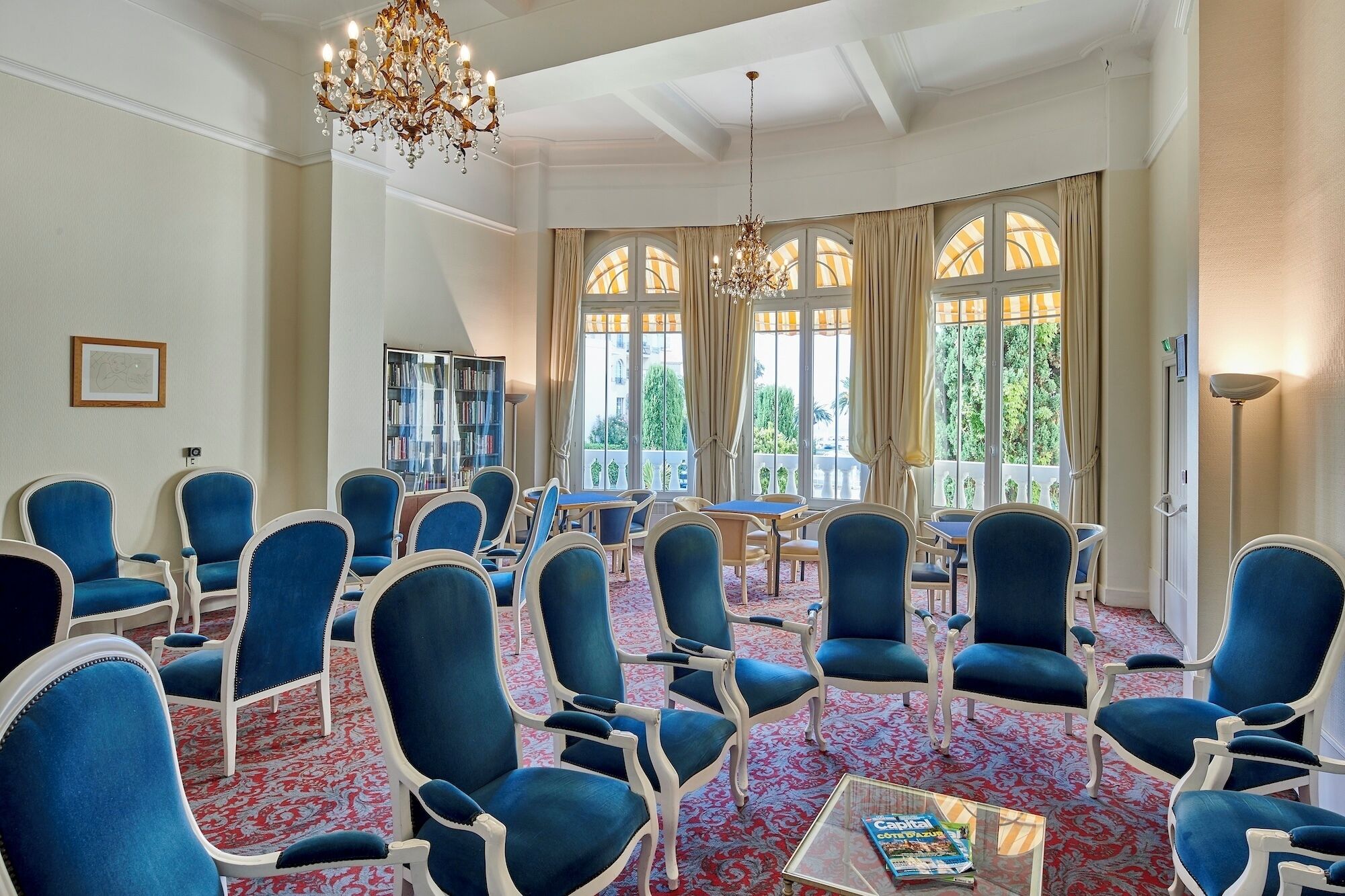 A room with blue upholstered chairs, a chandelier, a bookshelf, and large windows with curtains.
