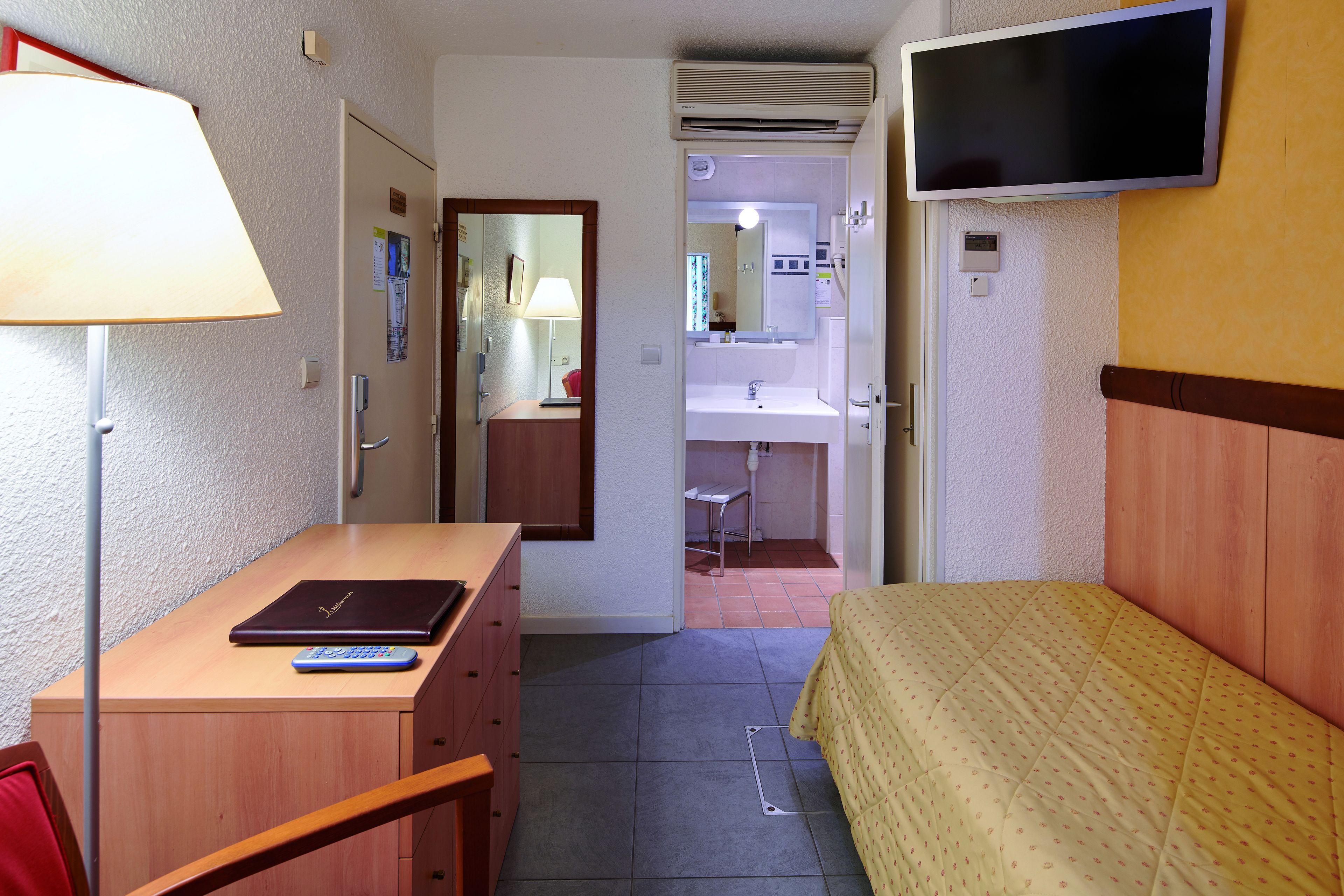 Habitació d'hotel senzilla amb un llit, escriptori amb un ordinador portàtil i un comandament a distància, televisió penjada a la paret i un bany visible a través d'una porta oberta.