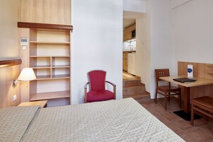 A hotel room with a bed, a red chair, a desk with a laptop, and a kitchenette in the background.