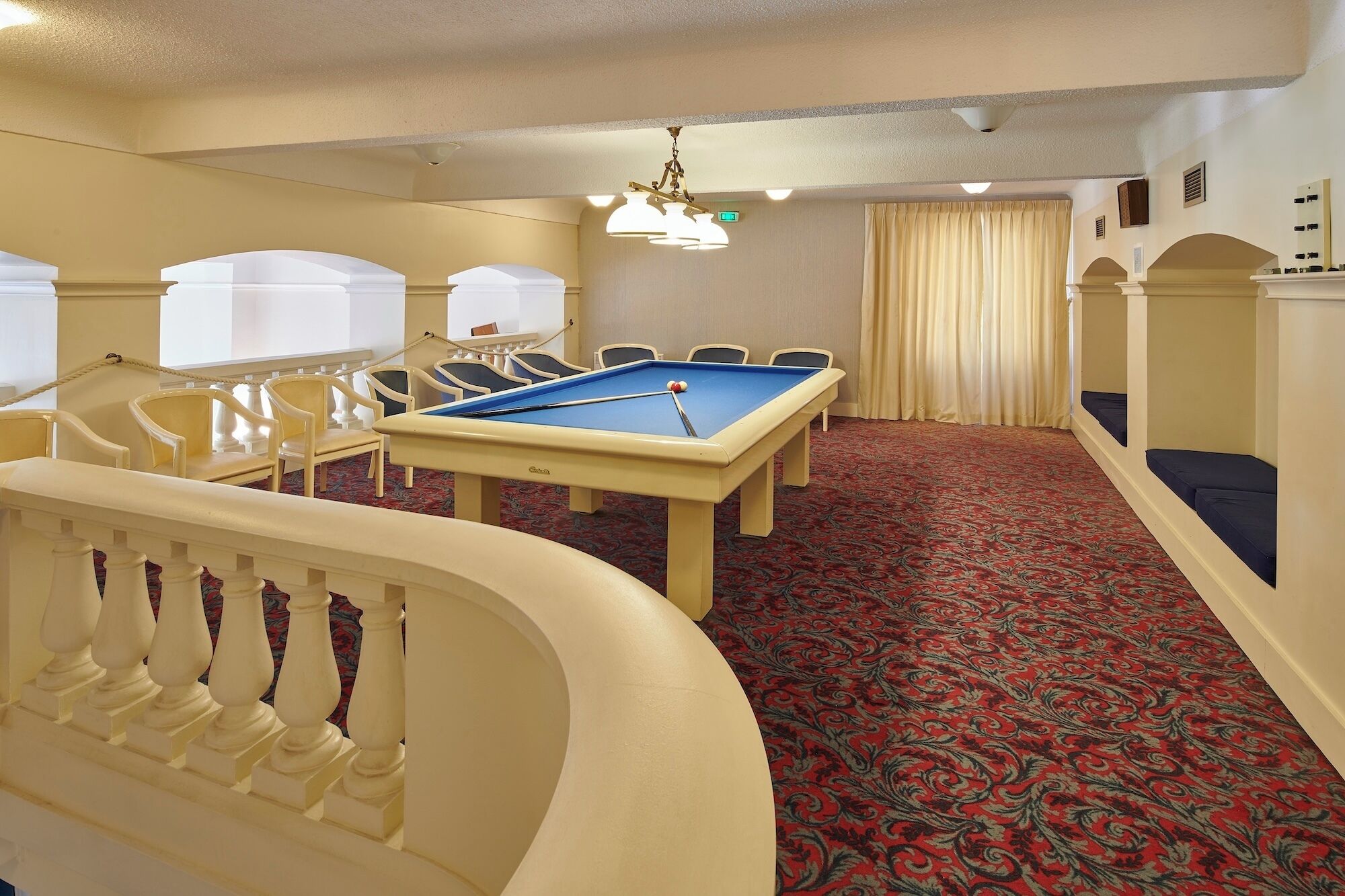 A pool table with chairs, a red patterned carpet, and a white railing.