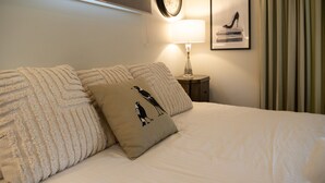 Ein ordentlich bezogenes Bett mit einem Kopfkissen mit Vogelmotiv, eine Nachttischlampe, eine Wanduhr und ein gerahmtes Bild über der Lampe.