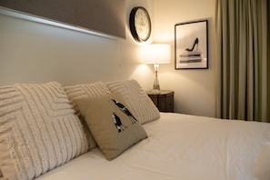 A neatly made bed with a pillow featuring a bird design, a bedside lamp, a clock on the wall, and a framed picture above the lamp.