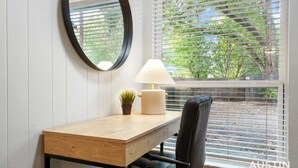 Une pièce avec un bureau en bois, une chaise noire, un miroir rond, une lampe et une fenêtre avec des stores.