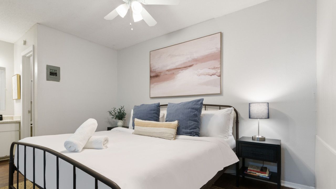 A neatly made bed with white linens and rolled towels, a bedside table with a lamp, and a framed abstract painting on the wall.