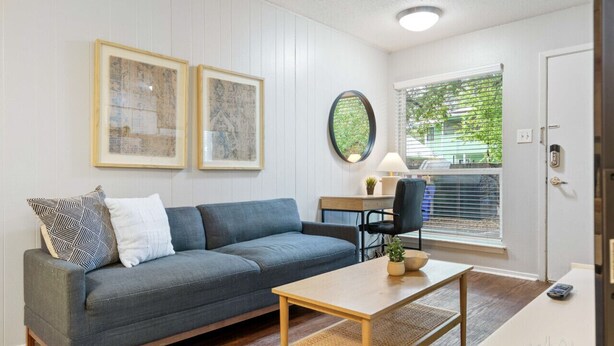 Un salon moderne avec un canapé gris, une table basse en bois et un bureau avec une chaise. Deux œuvres d’art encadrées ornent le mur, ainsi qu’un miroir rond et une fenêtre avec des stores.