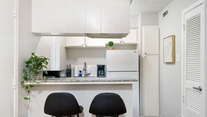 Une cuisine compacte avec des meubles blancs, un plan de travail, une machine à café et un réfrigérateur. On y trouve deux chaises noires et une plante en pot.
