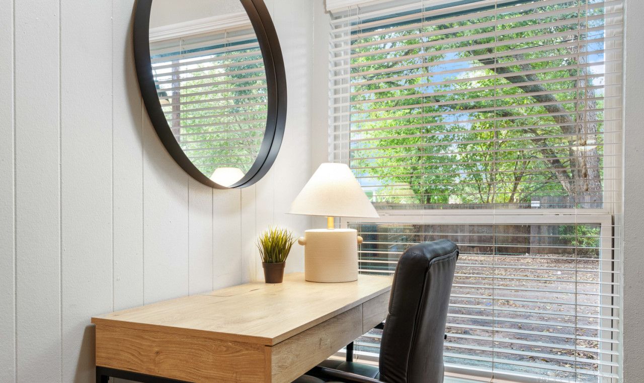 A room with a wooden desk, a black chair, a round mirror, a lamp, and a window with blinds.