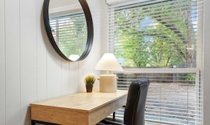Una habitación con un escritorio de madera, una silla negra, un espejo redondo, una lámpara y una ventana con persianas.