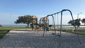 Une aire de jeux avec des balançoires et un toboggan, un mât pour drapeau, et une vue sur l’eau.