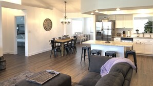 Un vaste salon comprenant un grand canapé gris, une table basse en bois et un coin repas avec une table et des chaises en bois. On aperçoit à l’arrière-plan une cuisine dotée de meubles blancs et d’appareils électroménagers en acier inoxydable.