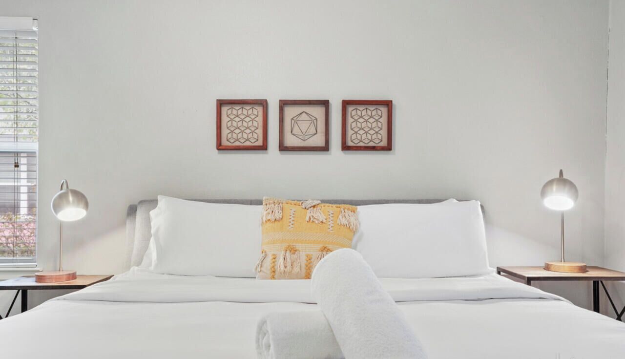 Ein ordentlich bezogenes Bett mit weißen Leinen, ein gelbes Kissen mit Muster und ein weißes Handtuch, das lässig über die Seite drapiert ist. Darüber hängen drei gerahmte Wandbilder mit geometrischen Formen.