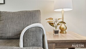 A grey sofa with a white throw blanket, a small table with a lamp and a plant, and a framed picture on the wall. - Kinney #4 xxxxxxxxxx (Austin)