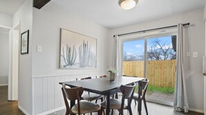 Un coin repas avec une table et des chaises, une grande fenêtre avec des rideaux et une œuvre d’art représentant un cactus accrochée au mur.