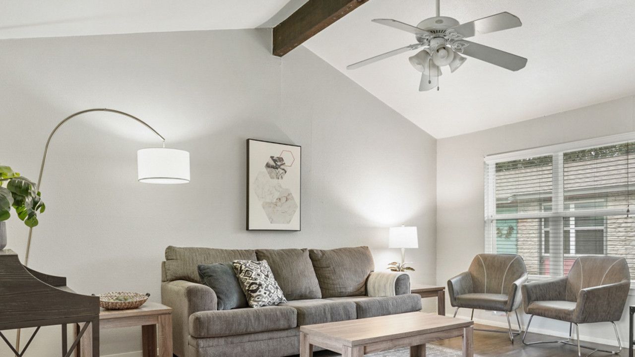 Ein modernes Wohnzimmer mit einem grauen Sofa, zwei Sesseln, einem Couchtisch, einer Stehlampe und einem Deckenventilator.