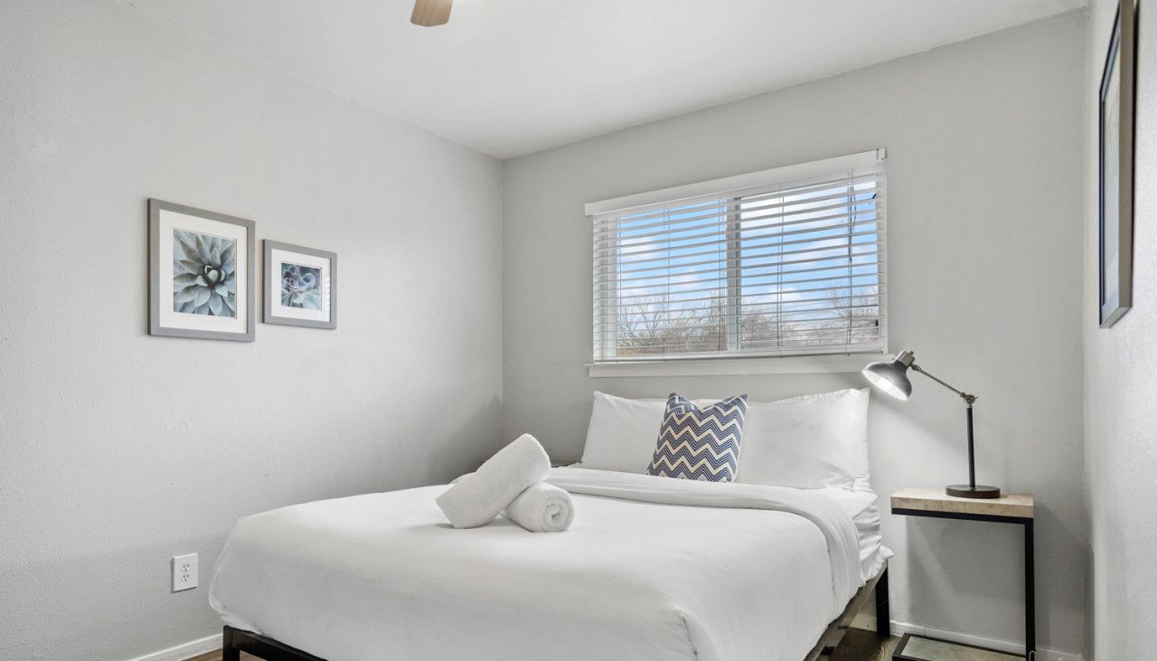 A bedroom with a bed, a nightstand, a lamp, a window with blinds, and two framed pictures on the wall.