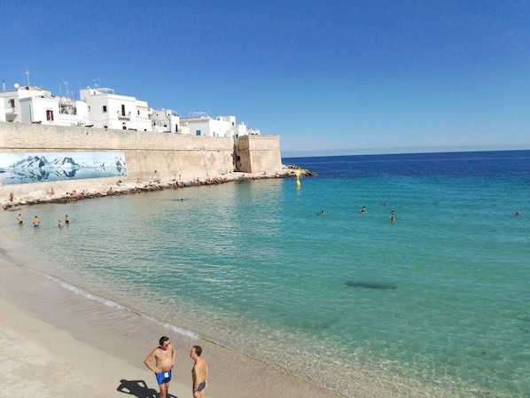 A beach with clear water, people swimming, and buildings in the background. - A step away from the sea - Apartment A / SELENE (Monopoli)