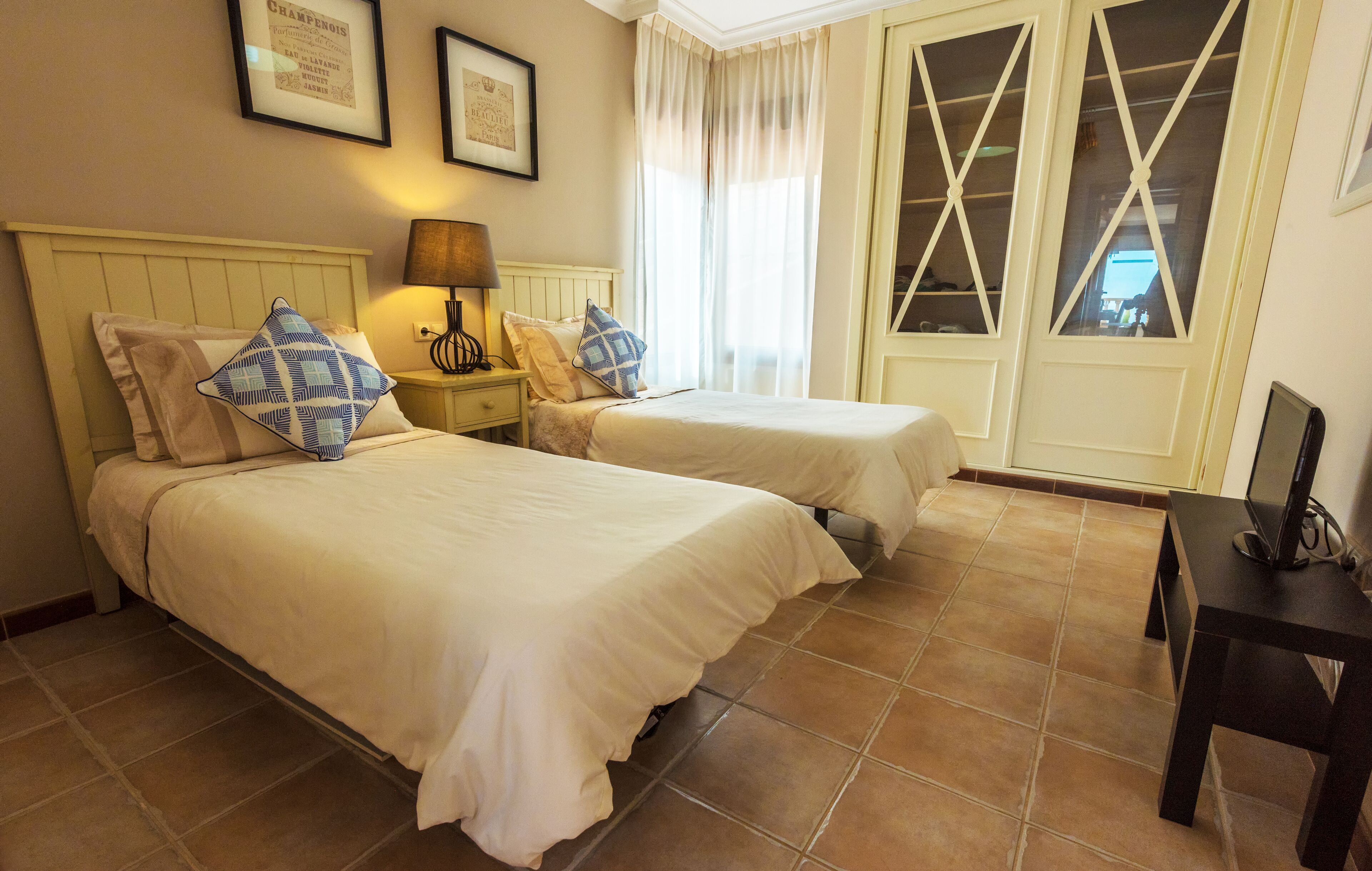 Une chambre d’hôtel avec deux lits, une table de chevet avec une lampe, un bureau avec un ordinateur et de grandes fenêtres avec des rideaux.