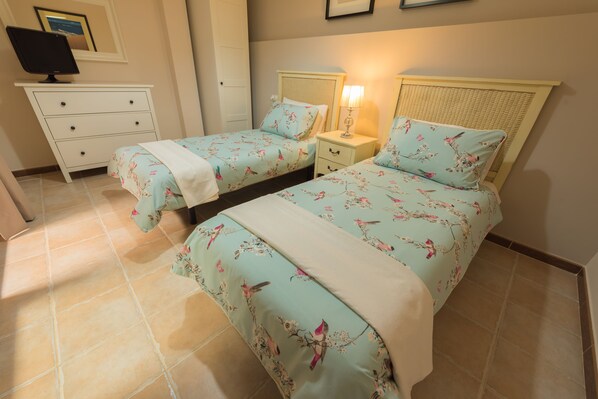 A bedroom with two single beds, a bedside table with a lamp, a dresser, and a television on a stand.