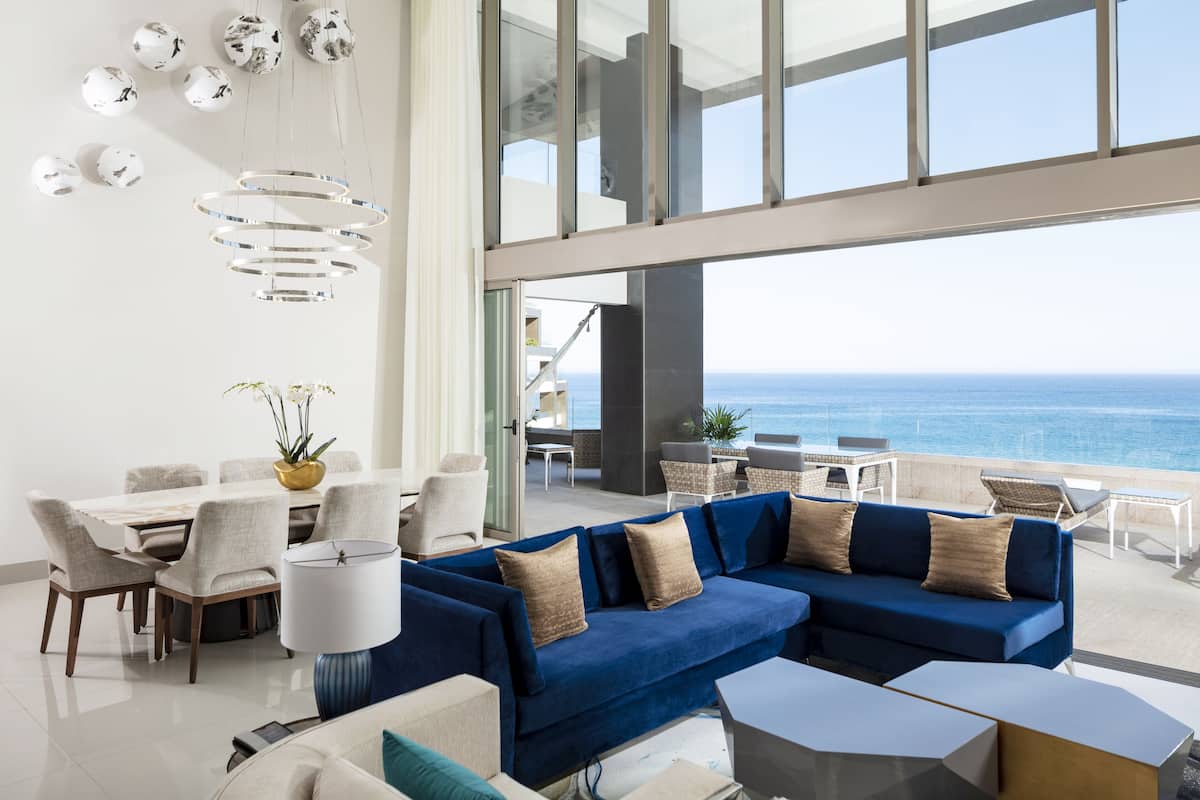 Ein modernes Wohnzimmer mit einem blauen Ecksofa, einem Essbereich mit weißem Tisch und Stühlen und Meerblick durch große Fenster.