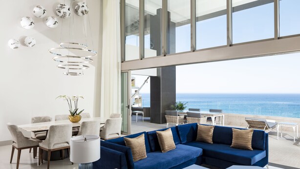 A modern living room with a blue sectional sofa, dining area with a white table and chairs, and a view of the ocean through large windows. - Garza Blanca Resort & Spa Los Cabos (Cabo San Lucas)