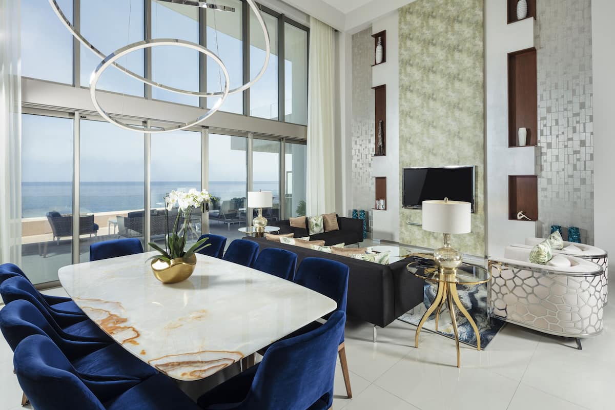Ein stilvoller Essbereich mit einem großen Marmortisch, blau gepolsterten Stühlen und Blick auf das Meer durch große Fenster.