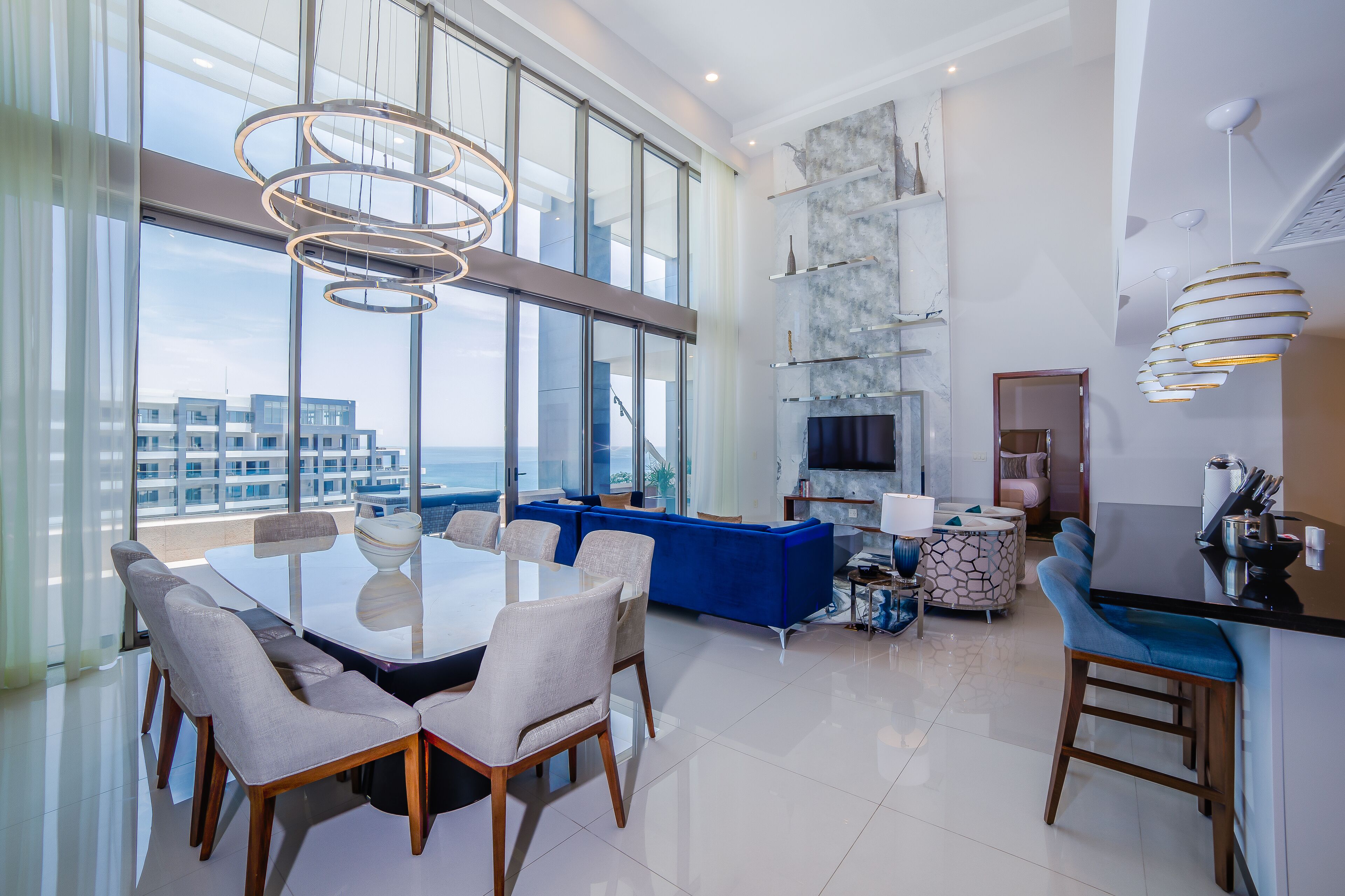 A modern living room with a large glass wall, a blue sofa, a dining table with chairs, and a kitchen area with a bar.