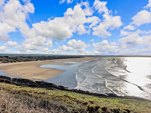 Un paisaje costero con una extensa playa de arena, un litoral rocoso y un cielo azul despejado con algunas nubes.