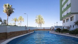 A swimming pool with a brick border, surrounded by palm trees and a building with a white and green facade. - Singular Stays Cullera Beach (Cullera)