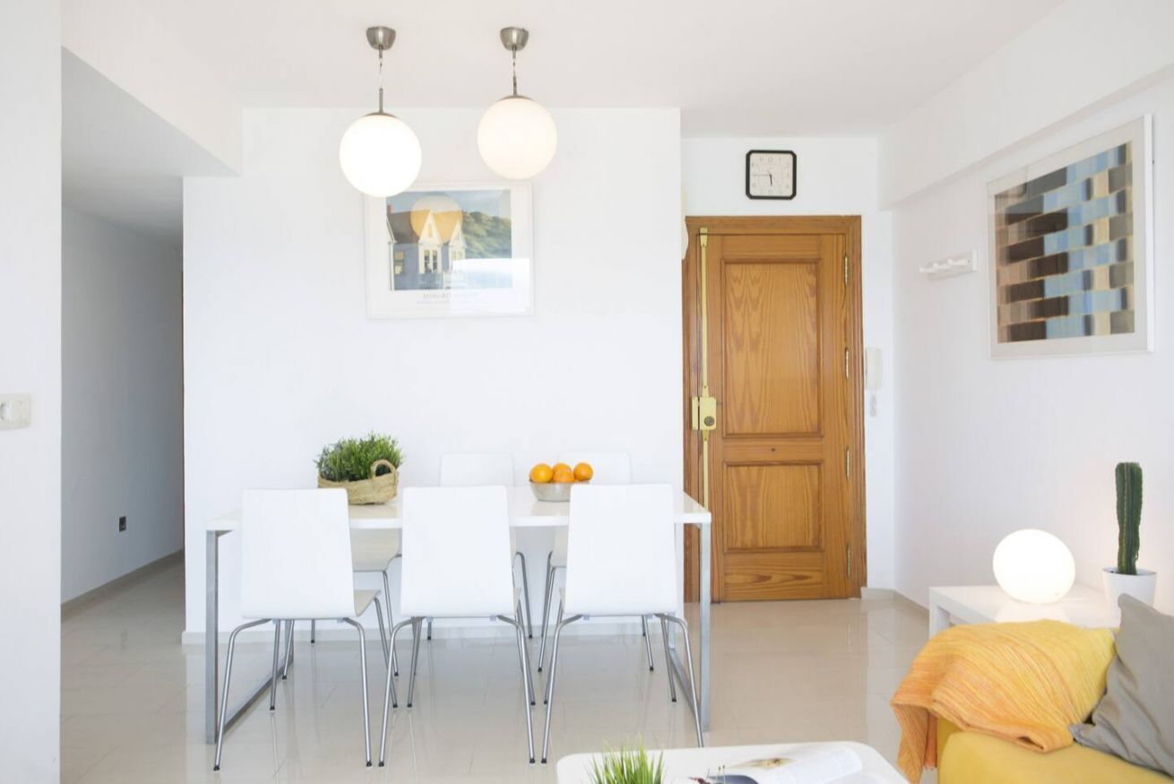 A modern kitchen with white cabinets, a wooden door, and a dining area with white chairs and a table.