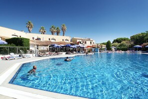 A swimming pool with lounge chairs and umbrellas, surrounded by buildings and palm trees. - Apartment in Saint-Loup amid Pine Trees (Agde)