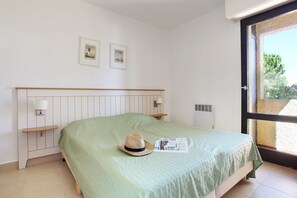 A bedroom with a bed, a nightstand, a lamp, two framed pictures on the wall, and a window with a view of greenery.