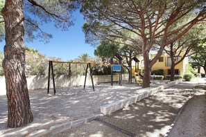 A playground with swings and a slide, surrounded by trees and a building in the background. - Apartment in Saint-Loup amid Pine Trees (Agde)