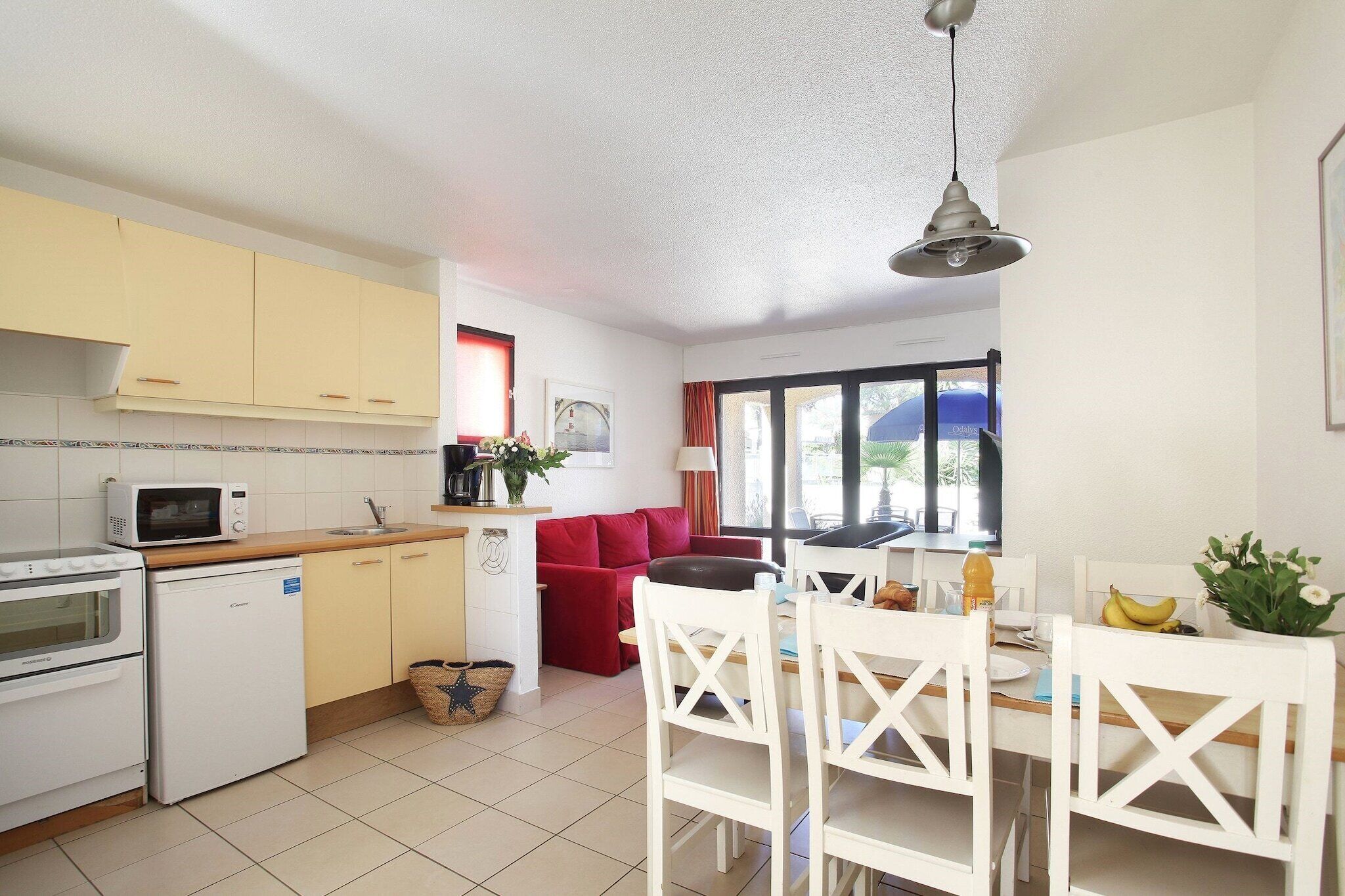 Een kleine keuken met een magnetron, oven en spoelbak. Er is een eethoek met witte stoelen en een tafel, en een rode bank op de achtergrond.