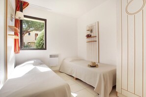 A room with two beds, a window with curtains, a radiator, and a wall-mounted shelf with a hat. - Apartment in Saint-Loup amid Pine Trees (Agde)