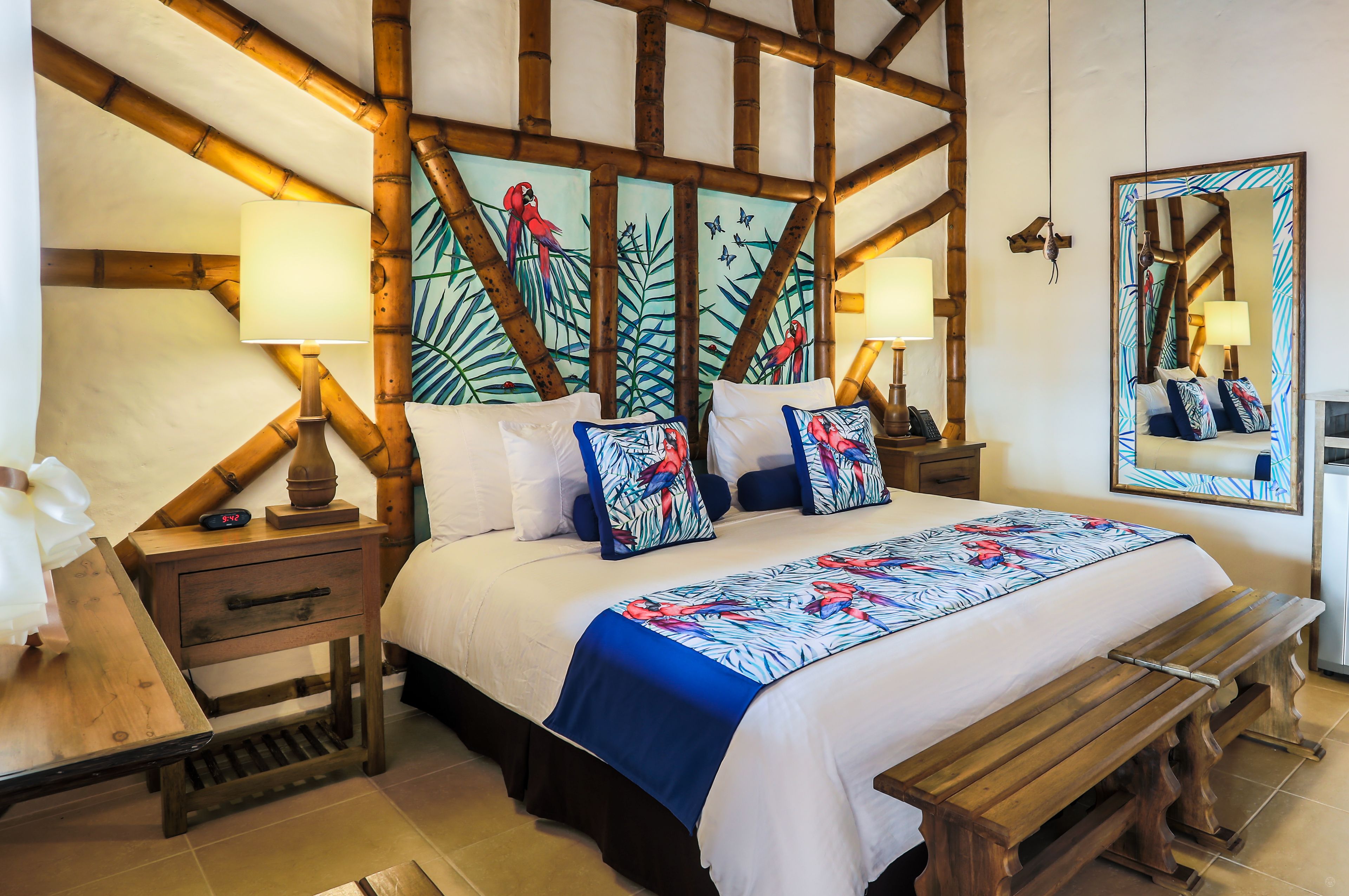 A hotel room with a large bed, wooden headboard, two bedside tables with lamps, a bench, and a mirror on the wall.