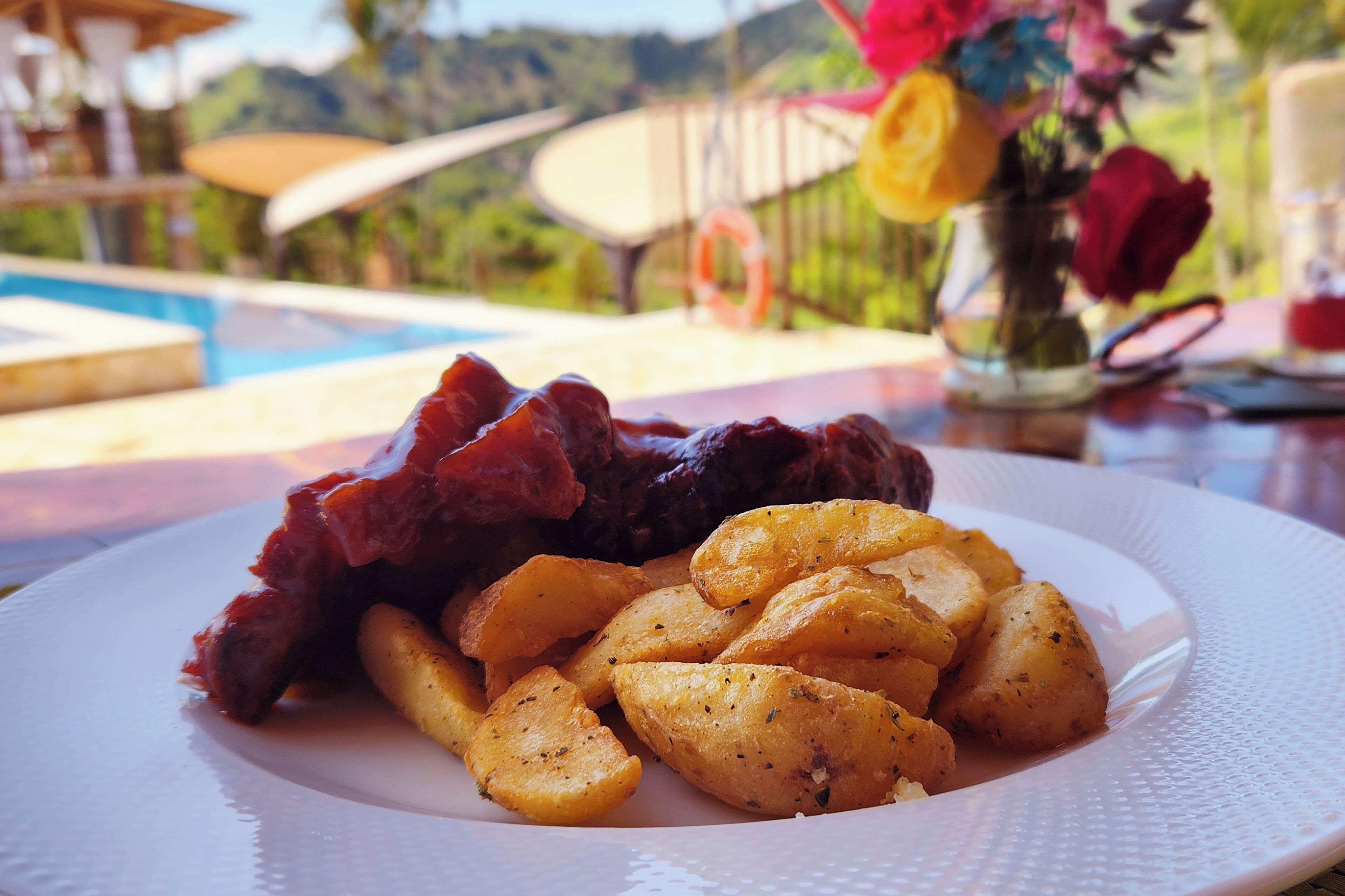 A plate of food with potatoes and meat, a pool, and a garden in the background.