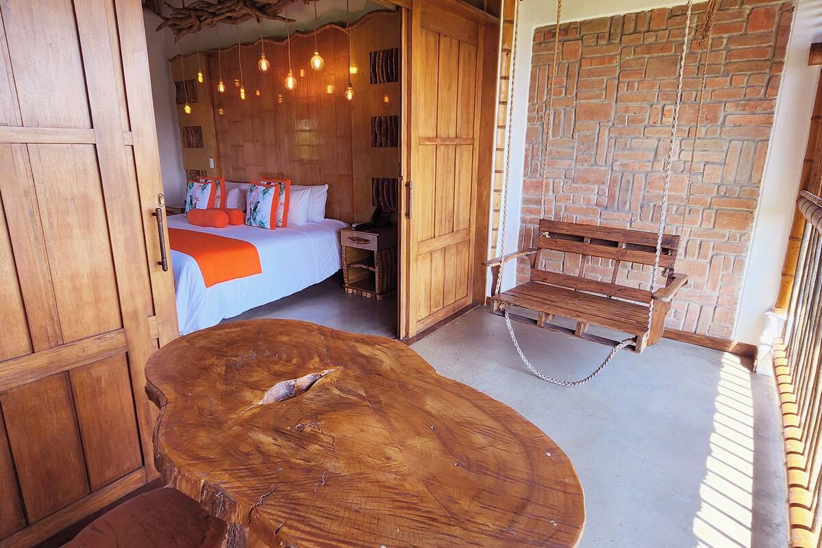 A wooden bed with orange bedding, a wooden bench, and a hanging swing in a room with a brick wall and wooden paneling.