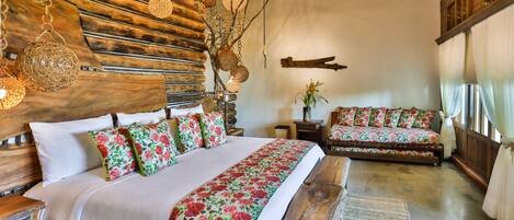 A bedroom with a wooden headboard, a large bed with floral bedding, a bench, and a small table with a plant.