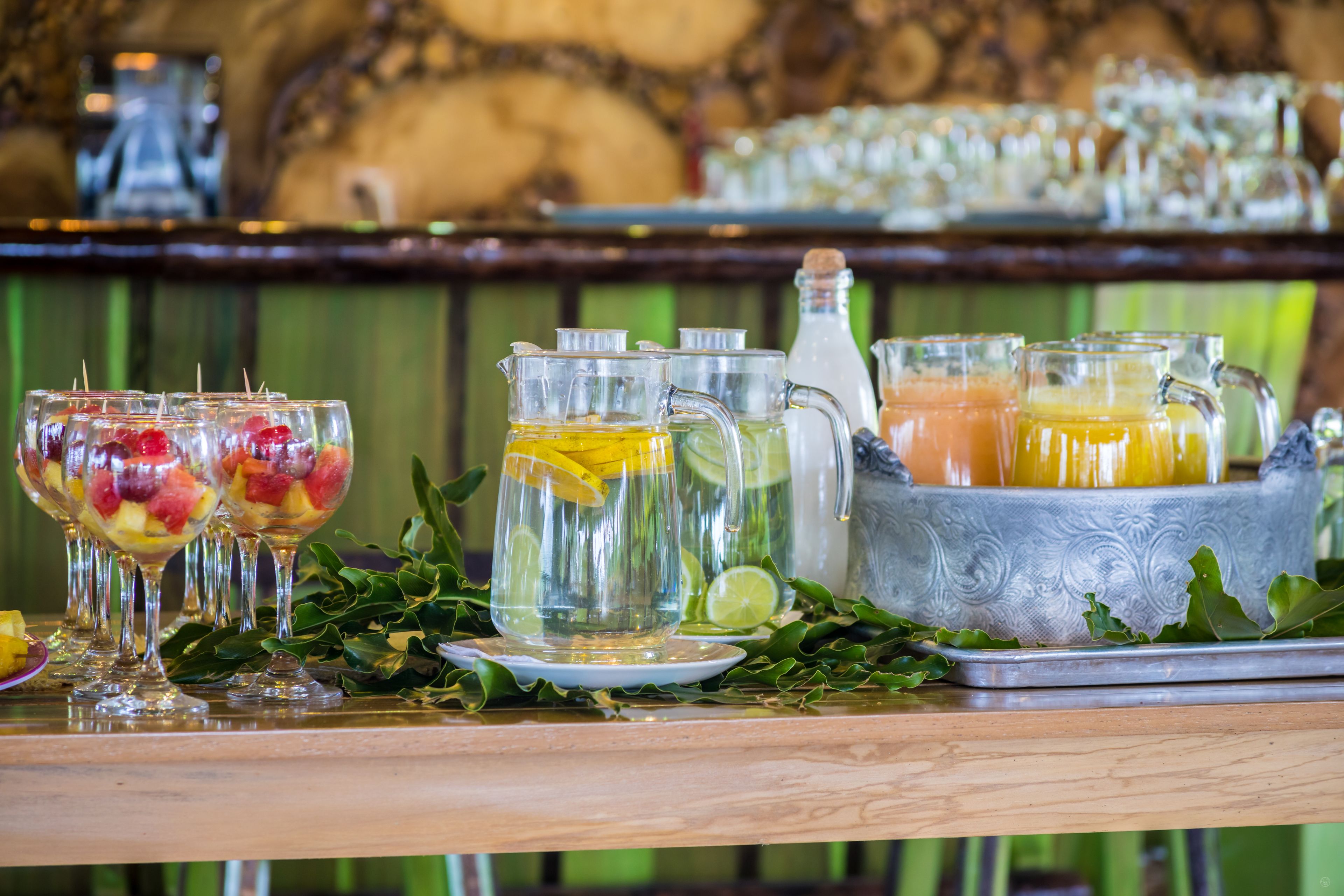 A bar area with various drinks, including fruit-infused beverages and pitchers, set on a wooden table with a decorative backdrop.