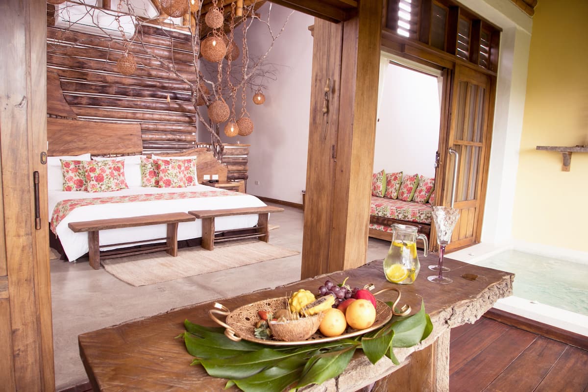 A wooden-framed bedroom with a bed, bench, and a breakfast table with fruits and a pitcher.