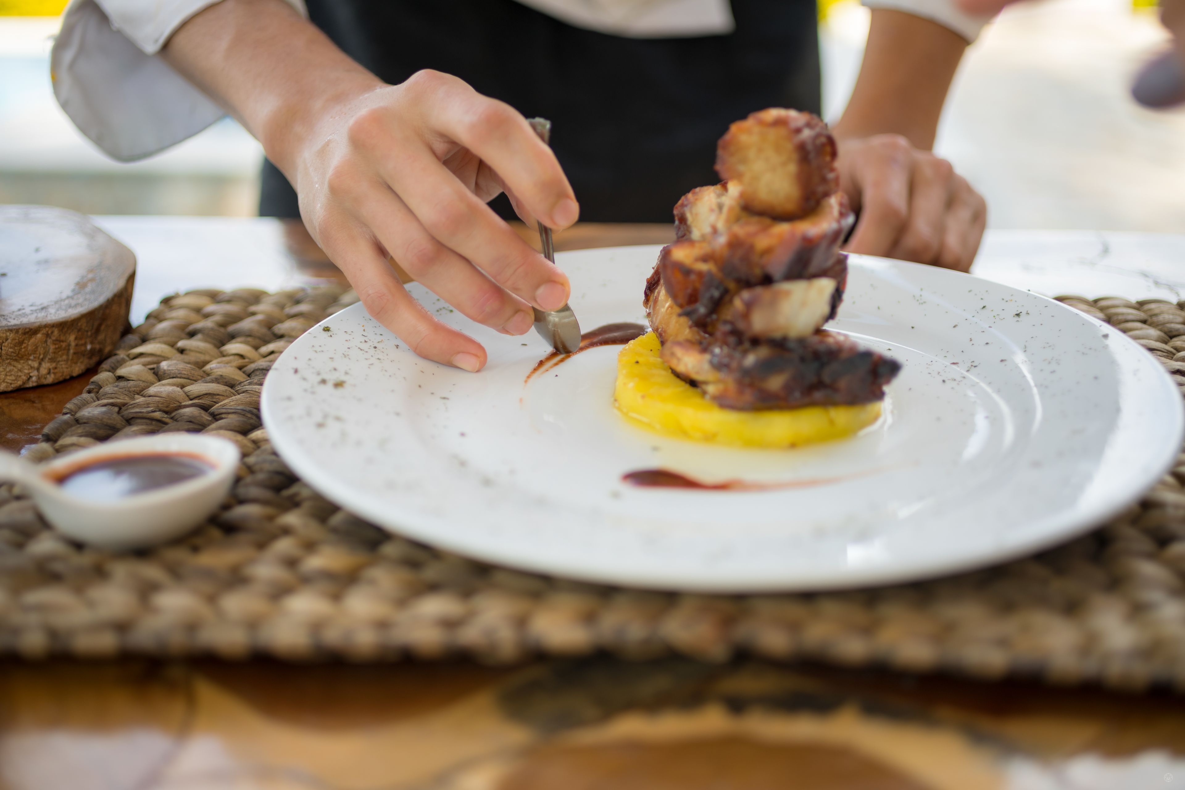 A person carving a piece of meat on a plate with a small bowl of sauce nearby.
