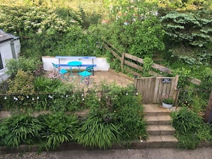 Un jardín con un portón de madera que dice "JULY COTTAGE", una pequeña mesa y sillas, y una variedad de plantas y flores.